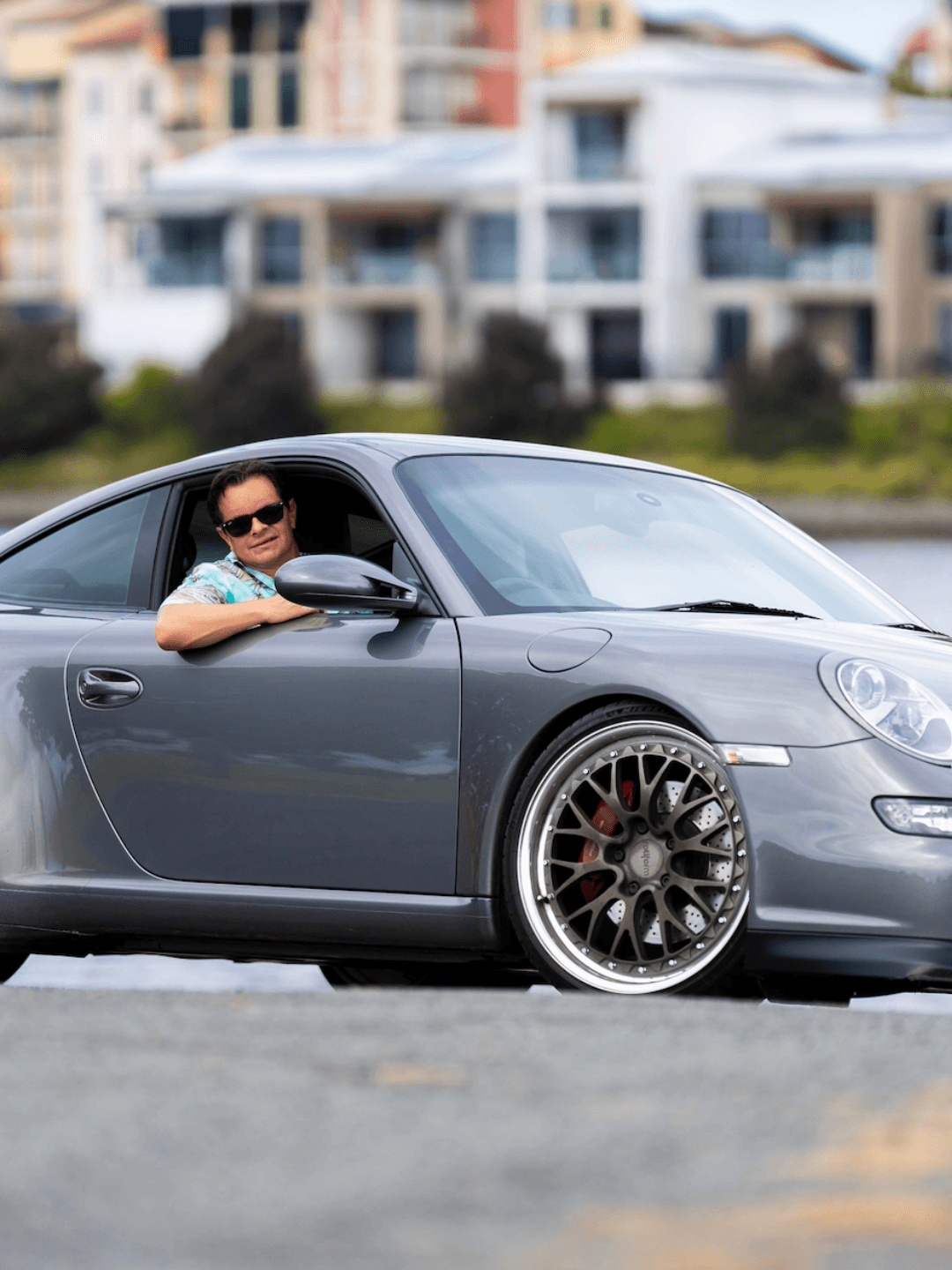 Greg Cassar leaning out of his grey Porsche 911 with aftermarket wheels, wearing sunglasses, with waterfront buildings in the background.